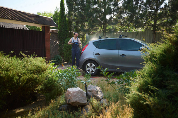 İş tulumlu genç adam, yüksek basınçlı temizleyici kullanıyor, arabasını arka bahçede yıkıyor. - Fotoğraf, Görsel