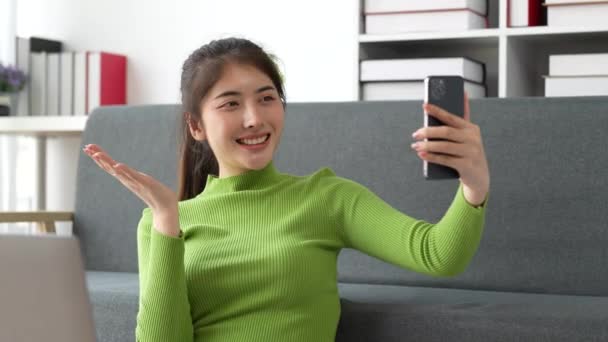 Nätti aasialainen nainen istuu sohvalla kotona nauttia videopuhelu tapaaminen ystävän kanssa. Nainen tekee sisältöä älypuhelimella kotona. Laadukas 4k kuvamateriaalia - Materiaali, video