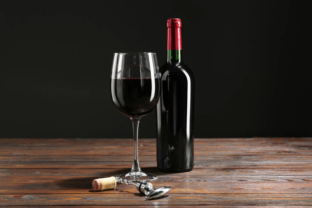 Corkscrew, glass and bottle of red wine on wooden table against black background - Foto, imagen