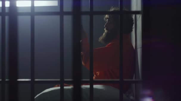 В'язень в помаранчевій формі сидить на ліжку, розмовляє по телефону в камері в'язниці з колючим вікном. Винний злочинець відбуває покарання у вигляді позбавлення волі. В'язень у СІЗО або виправній установі. - Кадри, відео