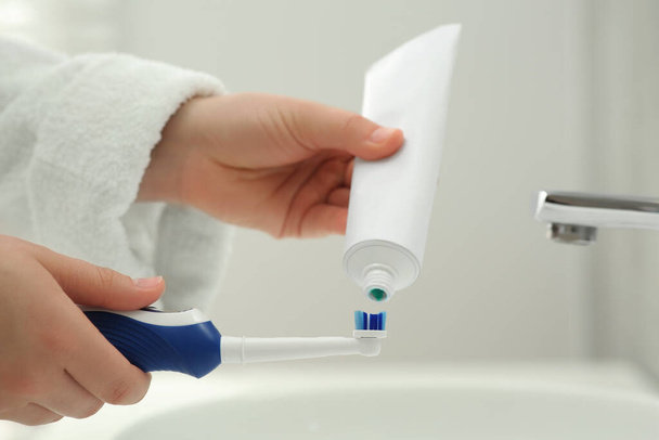 Vrouw knijpen tandpasta van buis op elektrische tandenborstel boven wastafel in de badkamer, close-up - Foto, afbeelding