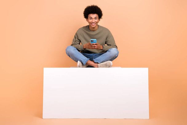 Full length photo of cheerful young man sit empty space podium use smart phone isolated on beige color background. - Foto, imagen