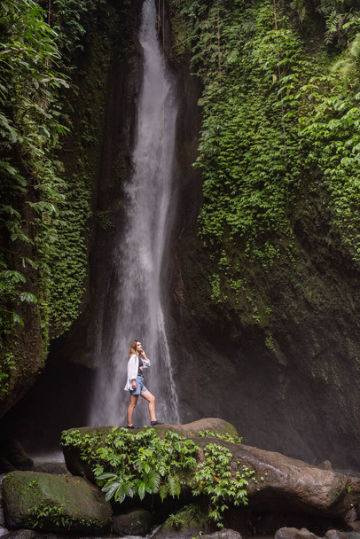 Travel lifestyle. Young traveler woman at waterfall in tropical forest. Leke Leke waterfall, Bali, Indonesia - Fotó, kép
