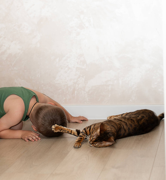 Un niño pequeño y hermoso de 4 años se está divirtiendo jugando con un gato de bengala leopardo rojo en el suelo en el interior de una casa. El gato doméstico yace en el suelo y juega con el bebé con su pata. - Foto, imagen