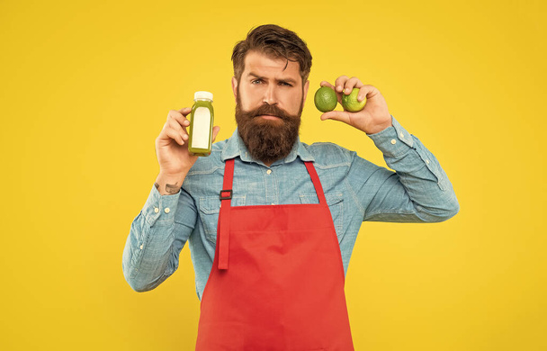 Serious man in apron holding limes and juice bottle yellow background, juice shopkeeper. - Valokuva, kuva
