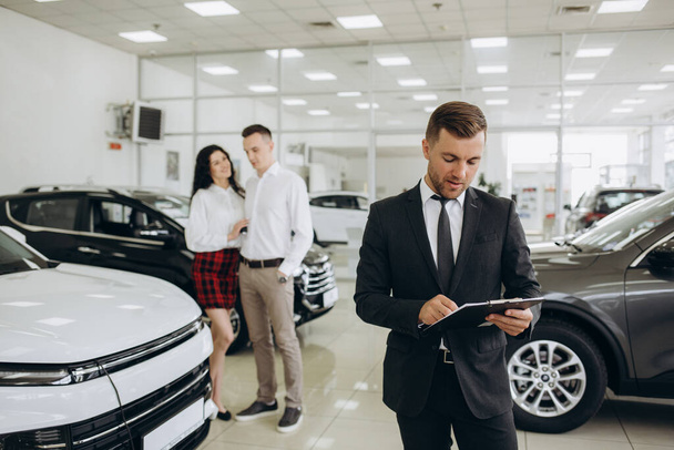 Closeup shot of successful confident smiling caucasian male shop assistant holding clipboard in formal clothes looking at camera at automobile car dealer shop. - Photo, image