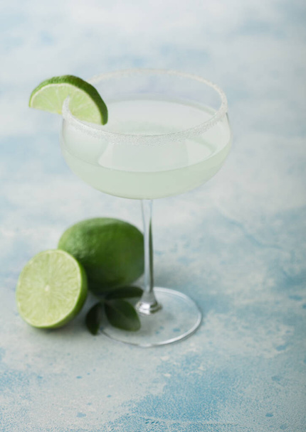 Crystal glass of Margarita cocktail with fresh limes on light blue table background. - Photo, Image
