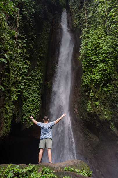 Back view of a man standing by waterfalls with arms outstretched. Man at waterfall raising his hands in feeling closer to nature. Man at the waterfall. Travel to Bali - Zdjęcie, obraz