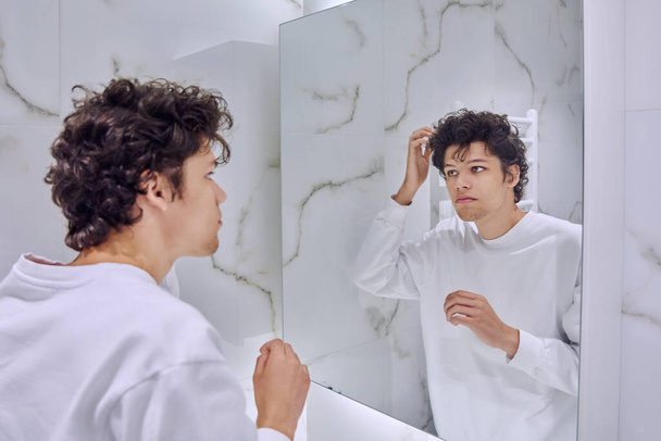 Young handsome male looking at himself in bathroom mirror, touching curly trendy hairstyle, admiring himself. Youth, fashion, beauty, hygiene, lifestyle, hair care, daily hair styling routine - Fotografie, Obrázek