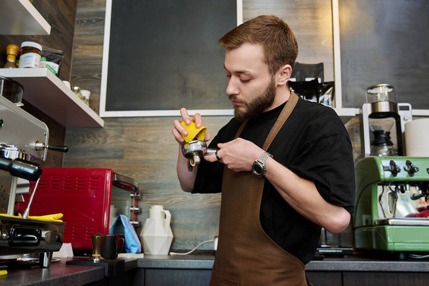 Male barista preparing coffee in coffee shop, bearded young man worker in an apron standing near coffee machine. Small business, food service occupation, coffee shop, staff, work concept - Zdjęcie, obraz