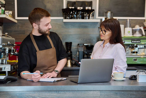 Colleagues partners, young male and mature woman talking working using laptop standing behind bar in coffee shop. Team, small business, work, staff, teamwork, entrepreneurship, partnership concept - 写真・画像