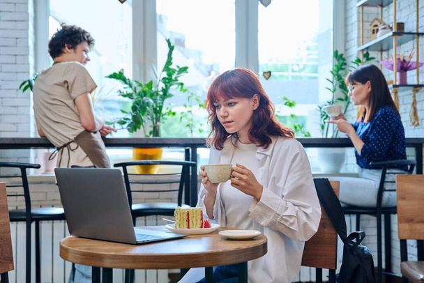 Female college student with laptop in cafe at table with cup of coffee and piece of cake. Internet online technology for leisure communication blogging learning chat, youth lifestyle concept - 写真・画像