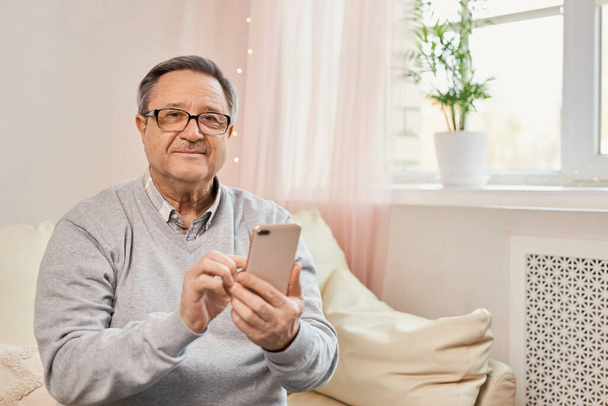 Menschen und Technologie. Porträt eines lächelnden erwachsenen Mannes, der sein Handy benutzt, Video anschaut oder SMS liest, zu Hause auf der Couch im Wohnzimmer sitzt. Älterer Mann nutzt moderne Technik - Foto, Bild