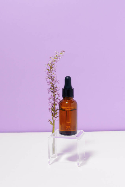 Vertical image of a glass transparent jar of facial serum with a flower on a purple background. Concept of face cosmetics, natural ingredients and beauty - Photo, Image