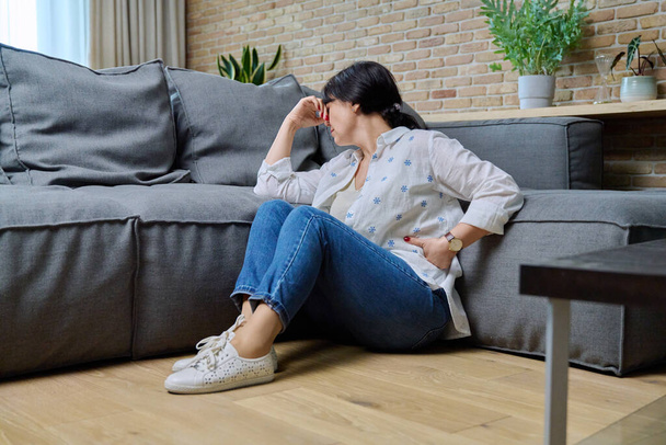 Mature woman suffering from physical pain, mental problems. 50s female sitting at home on floor in living room near sofa. Problems difficulties stress loneliness, age-related changes, health concept - 写真・画像