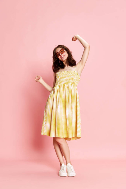 Full-length portrait of beautiful young woman in pretty yellow dress and sunglasses cheerfully dancing on pink studio background. Concept of human emotions, fashion, beauty, lifestyle, summer vibe, ad - Fotó, kép