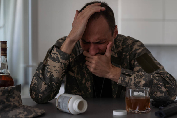 Ein Mann in Tarnkleidung leidet nach seiner Rückkehr aus der Armee an Depressionen. Er nimmt Alkohol und Betäubungstabletten. Er wird von schweren Erinnerungen gequält. Auf dem Tisch liegt ein Gewehr. Hochwertiges Foto - Foto, Bild