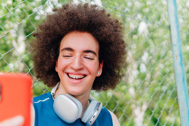 Glimlachende jongen met koptelefoon en sportieve kleding die een selfie maakt naast een urban basketbalveld - Foto, afbeelding