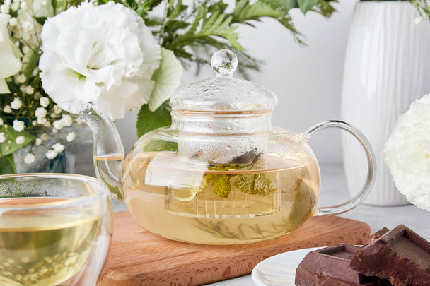 Esteettinen teeaika, teekannu, vihreä tee ja suklaa biofiliassa valkoisten kukkien joukossa. Naisellinen rauhallinen elämäntapa, kodikas kodin käsite. - Valokuva, kuva