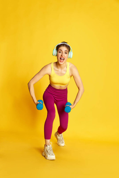 Full-length image of young sportive, slim woman in headphones training, doing exercises with dumbbells against yellow background. Concept of sport, fitness, body care, fashion, youth, lifestyle, ad - Foto, Imagen