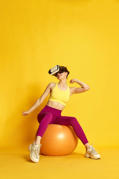 Full-length image of young slim woman in vr glasses sitting on fitness ball and training with dumbbells against yellow background. Concept of sport, fitness, body care, fashion, youth, lifestyle, ad - Foto, Imagen