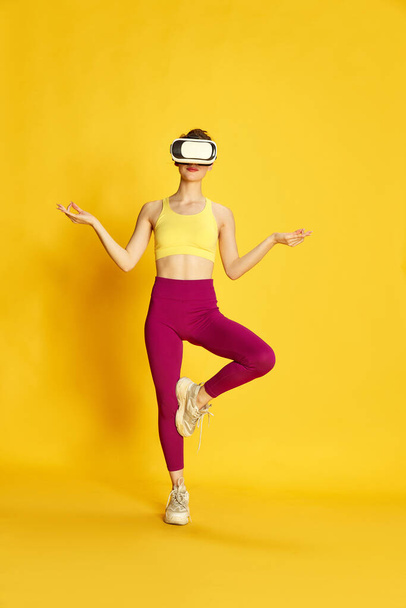 Full-length image of young slim woman wearing vr glases and training against yellow studio background. Yoga and meditation. Concept of sport, fitness, body care, fashion, youth, lifestyle, ad - Φωτογραφία, εικόνα