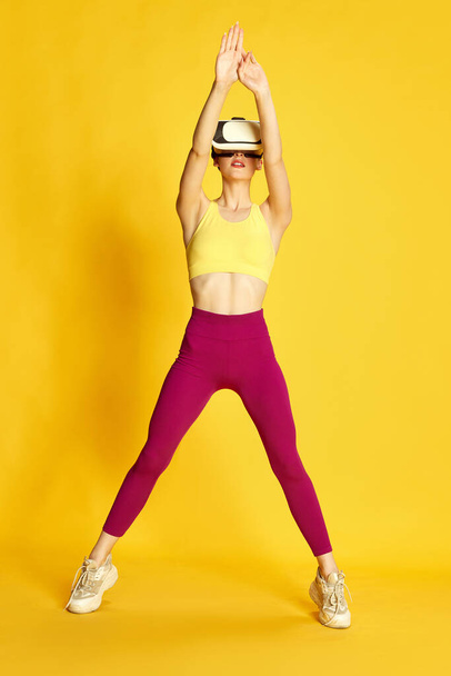 Full-length image of young slim woman wearing vr glases and training against yellow studio background. Stretching and yoga. Concept of sport, fitness, body care, fashion, youth, lifestyle, ad - Φωτογραφία, εικόνα