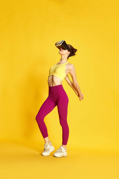 Full-length image of young sportive woman wearing vr glases and training against yellow studio background. Warming-up, stretching. Concept of sport, fitness, body care, fashion, youth, lifestyle, ad - Foto, immagini