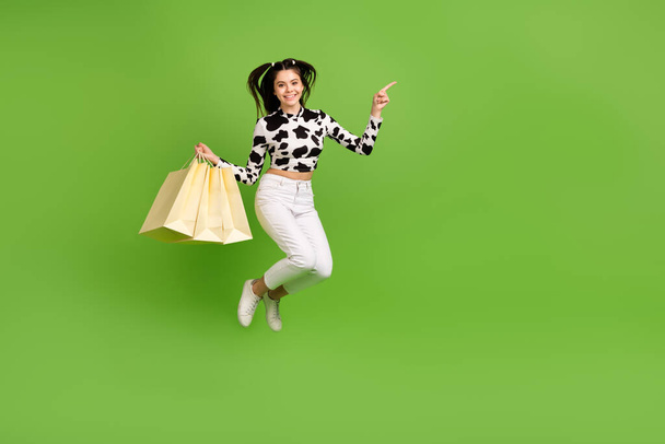 Full length photo of funny girl dressed cowskin top jumping high holding shoppers pointing empty space isolated green color background. - Fotografie, Obrázek