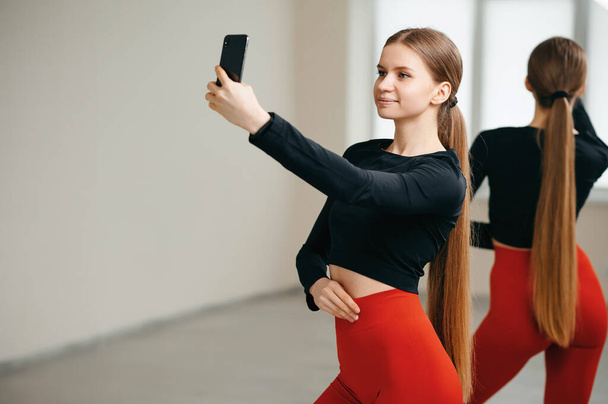 Smiling slender woman with long gathered hair in ponytail posing and making selfie on cellphone. Pretty female exercising in fitness center and using innovative technology. Training indoors. - Photo, image
