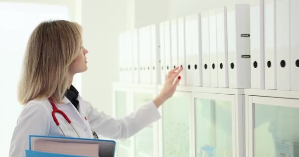 Γιατρός παίρνει λευκό φάκελο με δαχτυλίδια από το ράφι. Γυναίκα με ιατρική στολή αναζητά αρχειακό υλικό στο δωμάτιο της κλινικής - Πλάνα, βίντεο