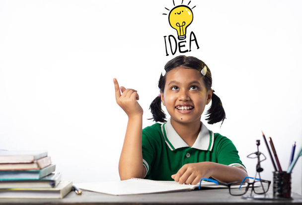 Красива індійська дитина, яка вчиться за навчальним столом з усміхненим обличчям, дивиться вгору на лампочку на білому фоні. - Фото, зображення