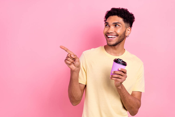 Photo of good mood guy with earrings dressed yellow t-shirt hold takeaway cup directing look empty space isolated on pink color background. - Foto, afbeelding