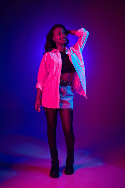 Full-length portrait of young, african, beautiful girl posing with smile against gradient blue purple studio background in neon light. Concept of human emotions, youth, fashion, lifestyle, ad - Photo, Image