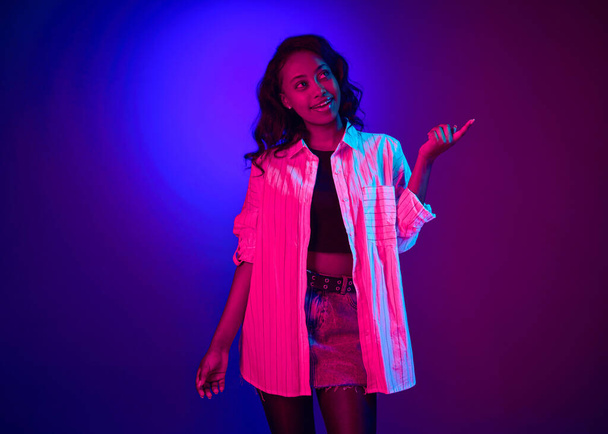 Portrait of young attractive african woman in white shirt posing with dreaming face over gradient blue purple studio background in neon light. Concept of human emotions, youth, fashion, lifestyle, ad - Foto, Bild