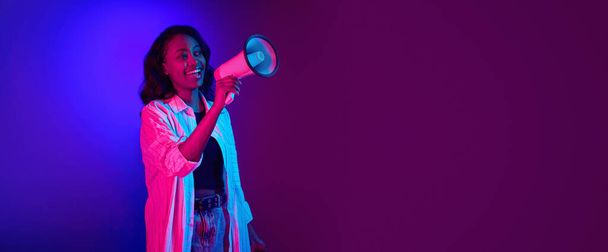 Portrait of young, smiling, african woman talking in megaphone against gradient blue purple studio background in neon light. News. Concept of human emotions, youth, fashion, lifestyle, ad. Banner - 写真・画像