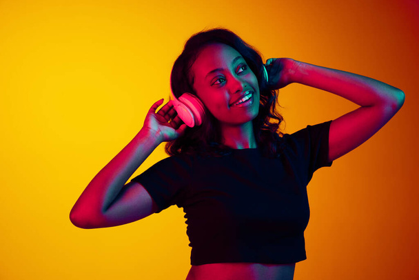 Portrét mladé krásné africké ženy, která poslouchá hudbu ve sluchátkách na pozadí žlutého studia v neonovém světle. Užijte si to. Pojem lidské emoce, mládež, móda, životní styl, reklama - Fotografie, Obrázek