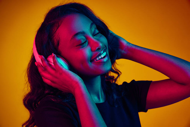 Portrait of young beautiful african woman listening to music in headphones, enjoying against yellow studio background in neon light. Concept of human emotions, youth, fashion, lifestyle, ad - Foto, immagini