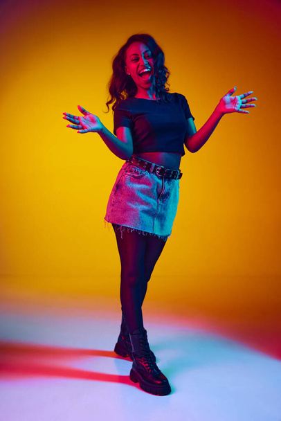 Full-length portrait of young beautiful african woman in stylish clothes cheerfully clapping hands against yellow studio background in neon light. Concept of human emotions, youth, fashion, lifestyle - Φωτογραφία, εικόνα