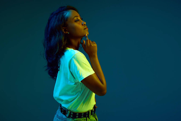 Portrait of young, african, beautiful woman in white t-shirt posing with thoughtful face against cyan, blue studio background in neon light. Concept of human emotions, youth, fashion, lifestyle, ad - Foto, Bild