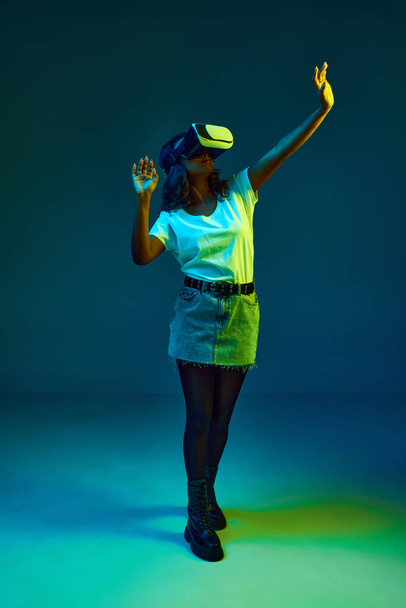 Portrait of attractive young, african woman in white t-shirt wearing vr glasses, posing on cyan, blue studio background in neon light. Innovations. Concept of emotions, youth, fashion, lifestyle, ad - Fotografie, Obrázek