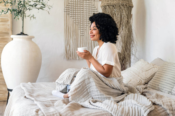 Африканская американка, покрытая одеялом, пьет горячий чай, чувствуя себя хорошо и улыбаясь. утренние выходные дома. концепция отдыха в праздничные дни. молодая женщина наслаждается чашкой кофе в постели - Фото, изображение