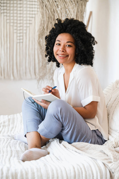 szczęśliwa afro amerykańska kobieta freelancer robi notatki w notatniku planowania podczas siedzenia w łóżku w domu. uśmiechnięta kobieta napisać pomysły inspiracji w pamiętniku - Zdjęcie, obraz