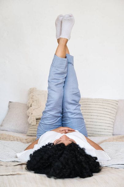 carefree african american woman lying on comfortable bed with legs raised up and rest at home bedroom. woman awake and relaxing in morning - Foto, imagen