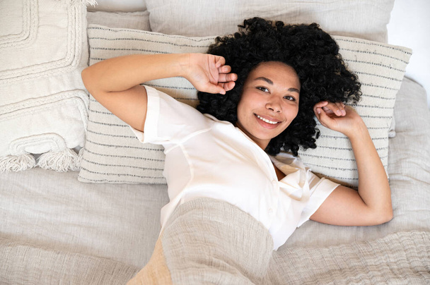 top view of smiling african american woman waking up after sleep and stretching with raised hands in cozy bedroom. positive female lying on bed with pillows and looking at camera - Photo, image