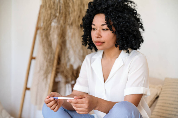 smutna, rozczarowana afro amerykańska kobieta trzyma test ciążowy siedząc sama na kanapie w domu. młoda samica ma dolegliwości, problemy zdrowotne lub niepłodność - Zdjęcie, obraz