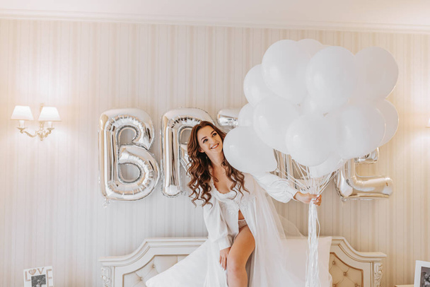 young curly girl with long hair in luxurious room standing on bed in white open dress holding many gel inflatable balls in hands and preparing for wedding ceremony - Fotografie, Obrázek