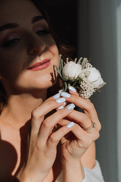 the groom's boutonniere is arranged from rose flowers in the hands of the bride with a French manicure in the wonderful natural light from the window - Photo, Image