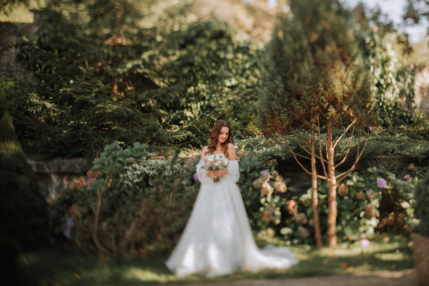 jonge en mooie bruid met lang bruin haar in een trouwjurk buiten met een bruiloft boeket bloemen. Portret op de TS-E lens - Foto, afbeelding