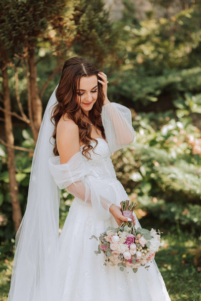 young and beautiful bride with long brown hair in a wedding dress outdoors with a wedding bouquet of flowers. Portrait of a girl in a white dress - Photo, Image